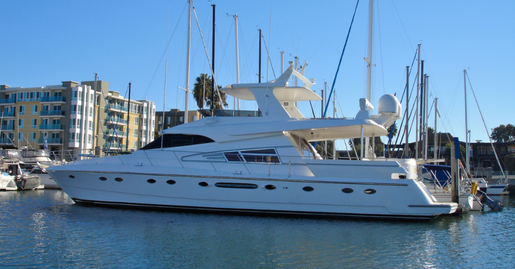 White luxury motor yacht moored in a marina with sailboats and modern buildings in the background.