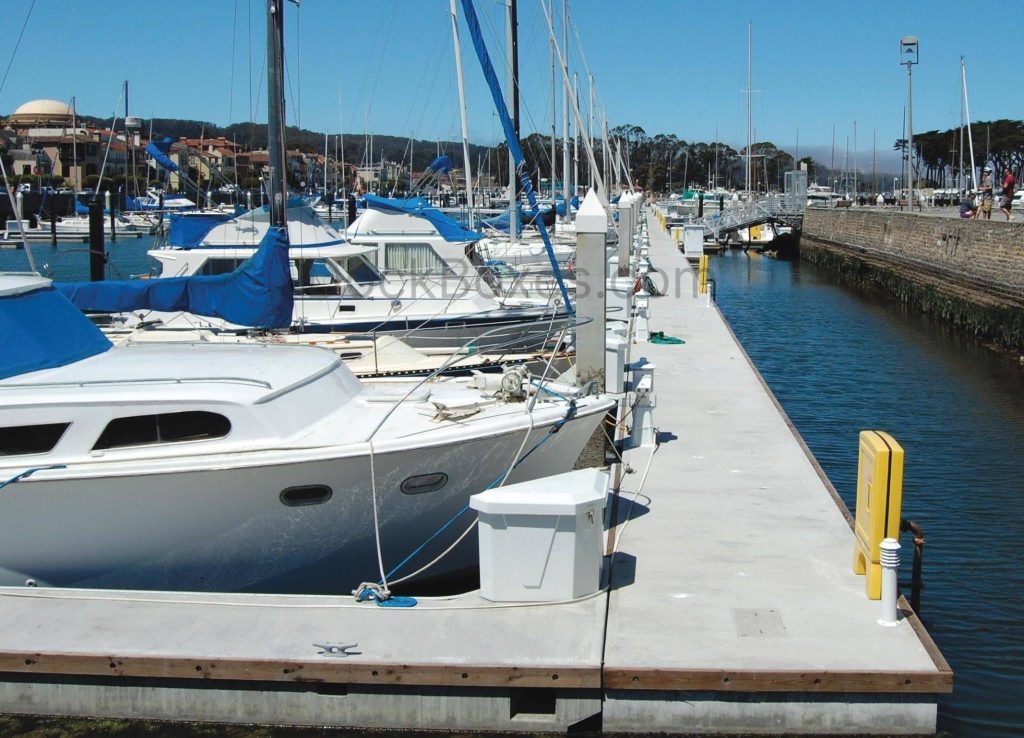 Marina with white motor yachts tied to a pier under a clear blue sky, calm water on the right side, and a walkway along the harbor edge.
