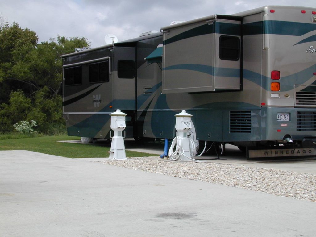 Two large motorhomes parked side by side on a paved lot with electrical pedestals and hoses in front, a grassy area and trees in the background.