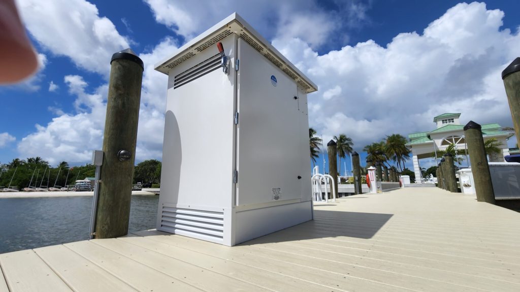 White outdoor electrical cabinet on a wooden marina dock with palm trees and a blue sky in the background.