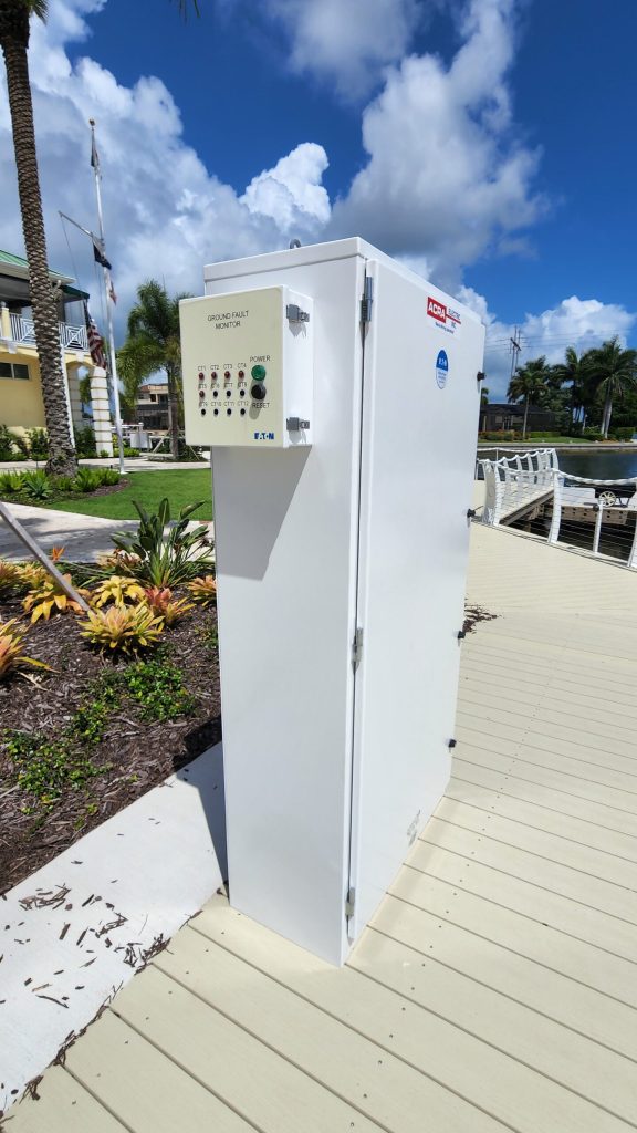 White outdoor electrical cabinet with a ground fault monitor panel, mounted on a deck near palm trees and a marina backdrop.