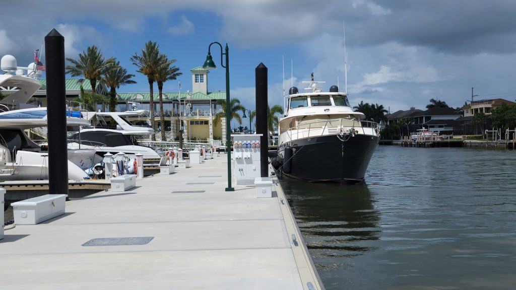 Marina with a large motor yacht docked at the pier, smaller boats along the left, palm trees and waterfront buildings in the background.