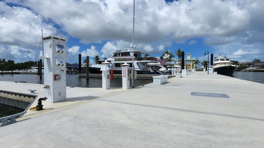 Marina with large white yachts docked along a concrete walkway, fueling pumps nearby and an American flag flying by a boat.