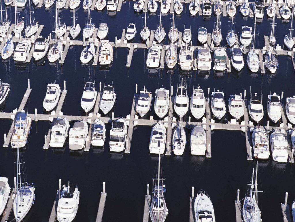 Aerial view of a crowded marina with numerous white boats docked along piers in calm blue water, viewed top-down.
