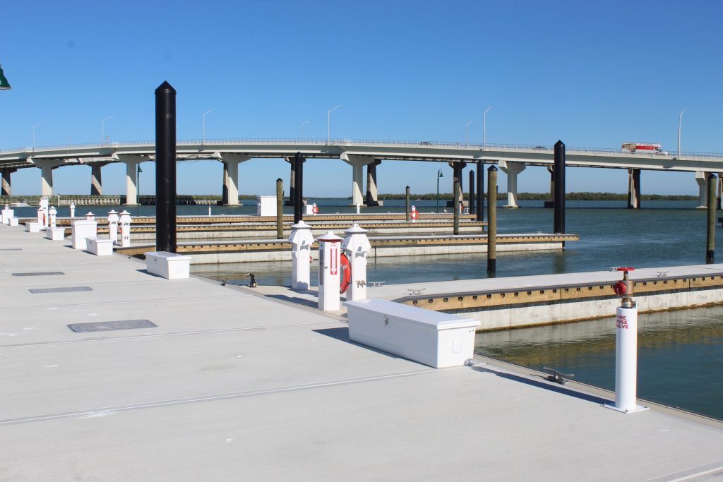 Wide view of a marina with docks, pilings, and a distant elevated bridge against a clear blue sky.