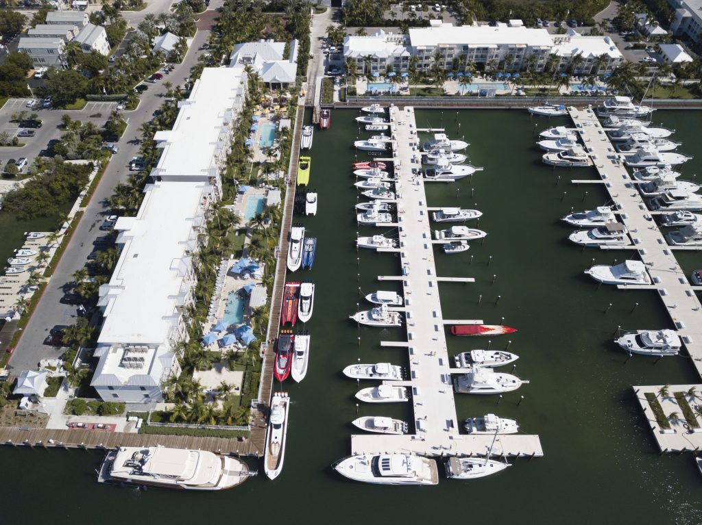 Aerial view of a marina with numerous white boats docked along parallel piers beside hotel-style buildings and palm trees.
