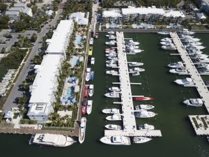 Aerial view of a marina with numerous white boats docked along parallel piers beside hotel-style buildings and palm trees.