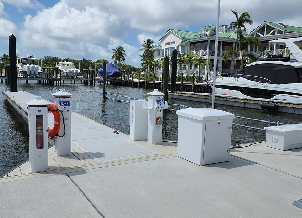 Marina dock with boat fuel pumps and life ring, calm water and yachts moored along slips under palm trees and pastel houses in the background.