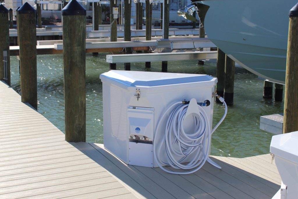 White utility box with a coiled hose on a wooden dock beside a moored boat and pilings at a marina, water in the background