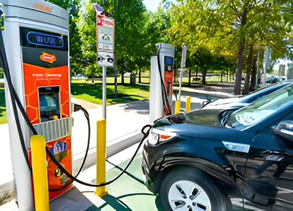 Electric vehicle charging station with a black car plugged in; orange charging unit and yellow bollards in a parking lot, trees in the background.