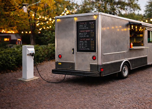 Silver food truck parked on gravel at dusk with string lights overhead and a chalkboard menu on the back panel