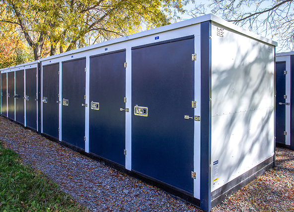 Row of blue outdoor storage lockers with silver trim along a gravel path, with autumn trees in the background.
