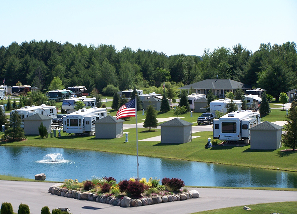 RV park with numerous touring trailers around a small pond with a fountain, flagpole, and green landscaping near trees and houses.