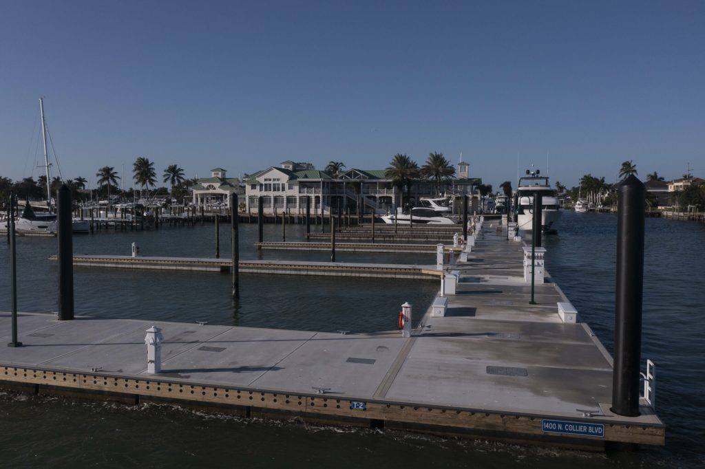 Wide view of a harbor with boats docked at wooden piers and a pale green-roofed clubhouse against a clear blue sky.